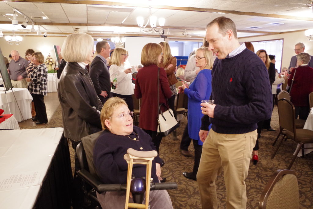 A female and male guest talking. The female guest is seated in a wheelchair with crutches wearing a black sweater, the male guest is standing wearing a blue sweater.
