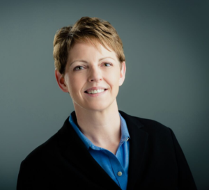 Deb is smiling with short blonde hair wearing a blue collared shirt and black blazer.