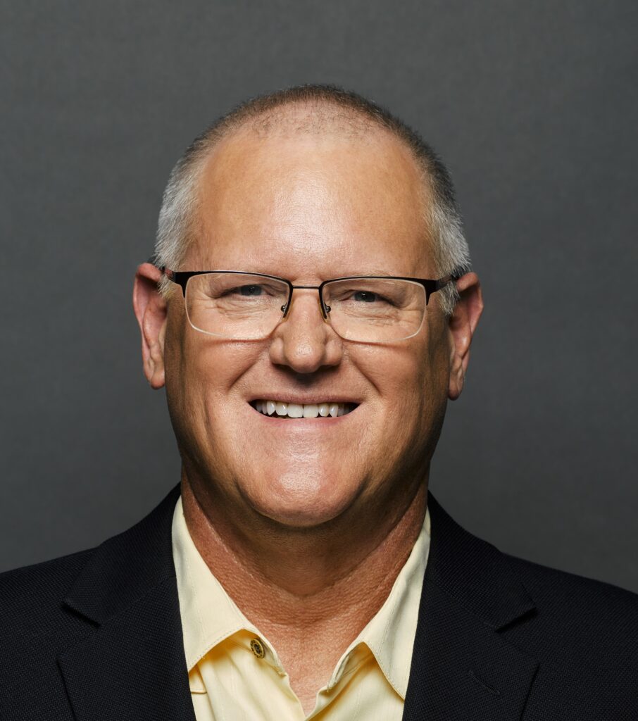 A portrait of Ken smiling. Ken is wearing glasses, a soft yellow button up shirt with a black suit jacket.